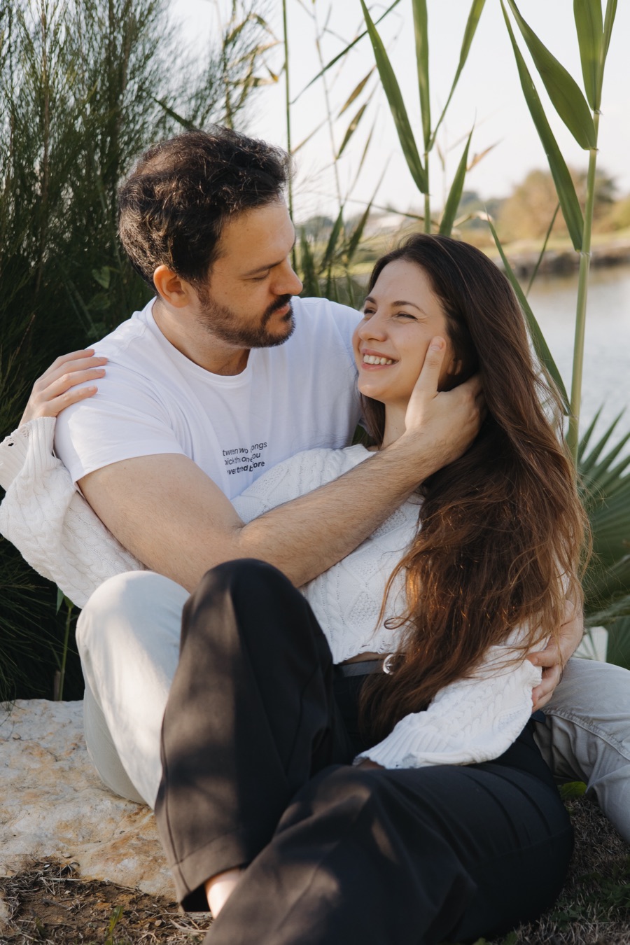 זוג בסוודרים לבנים יושב ליד המים, היא נוגעת בפנים שלו