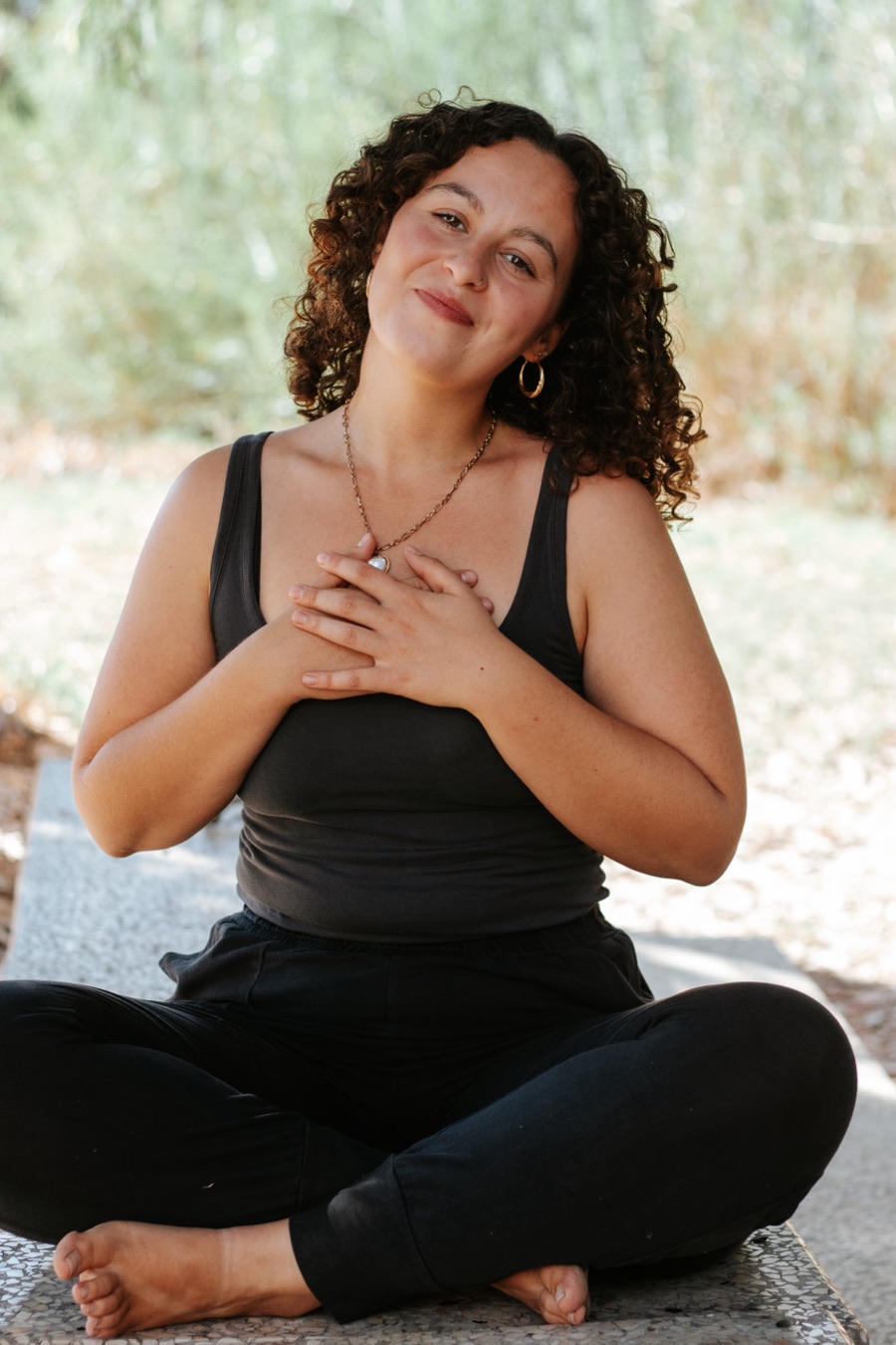 אישה עם תלתלים יושבת בחוץ, יד על הלב ומבט חם למצלמה